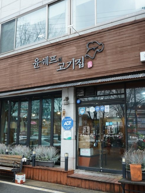 가게 바깥(세로) — 갈색 나무 간판에 은색 입체 글자 «윤쉐프의 고기집», 요리사 모자 선 로고, 분홍 «食» 도장. 유리 출입문에 같은 글씨·로고, ADT 보안 스티커, 왼쪽 접이식 창, 화분과 나무 벤치. 왼쪽 끝에 흰 승용차 뒷부분(번호판 일부가 보인다). 출입문 유리 안쪽에 흐린 사람 모습 하나(얼굴은 알아볼 수 없다)