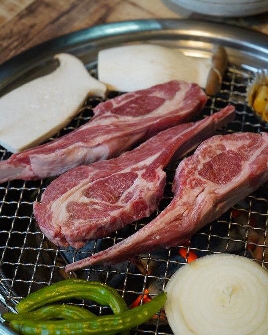 숯불 석쇠 위에 올린 생양갈비 세 대와 새송이버섯·양파·풋고추. 위쪽에 반찬 그릇