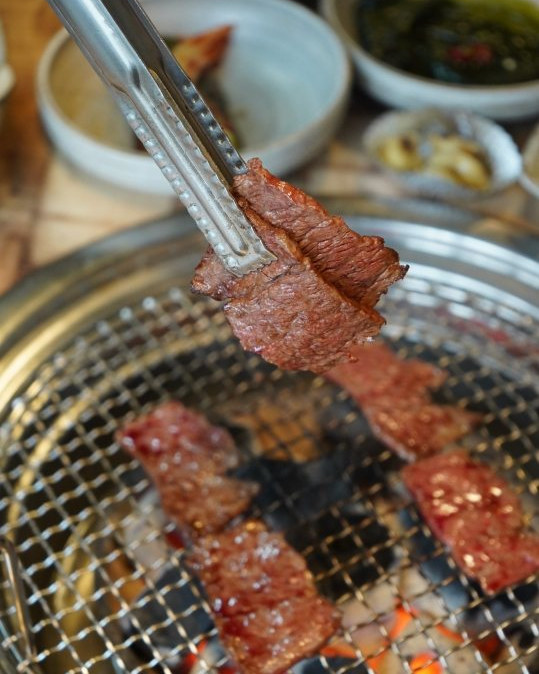 집게로 든 구운 고기 한 점, 석쇠 위 고기, 뒤로 밑반찬 그릇
